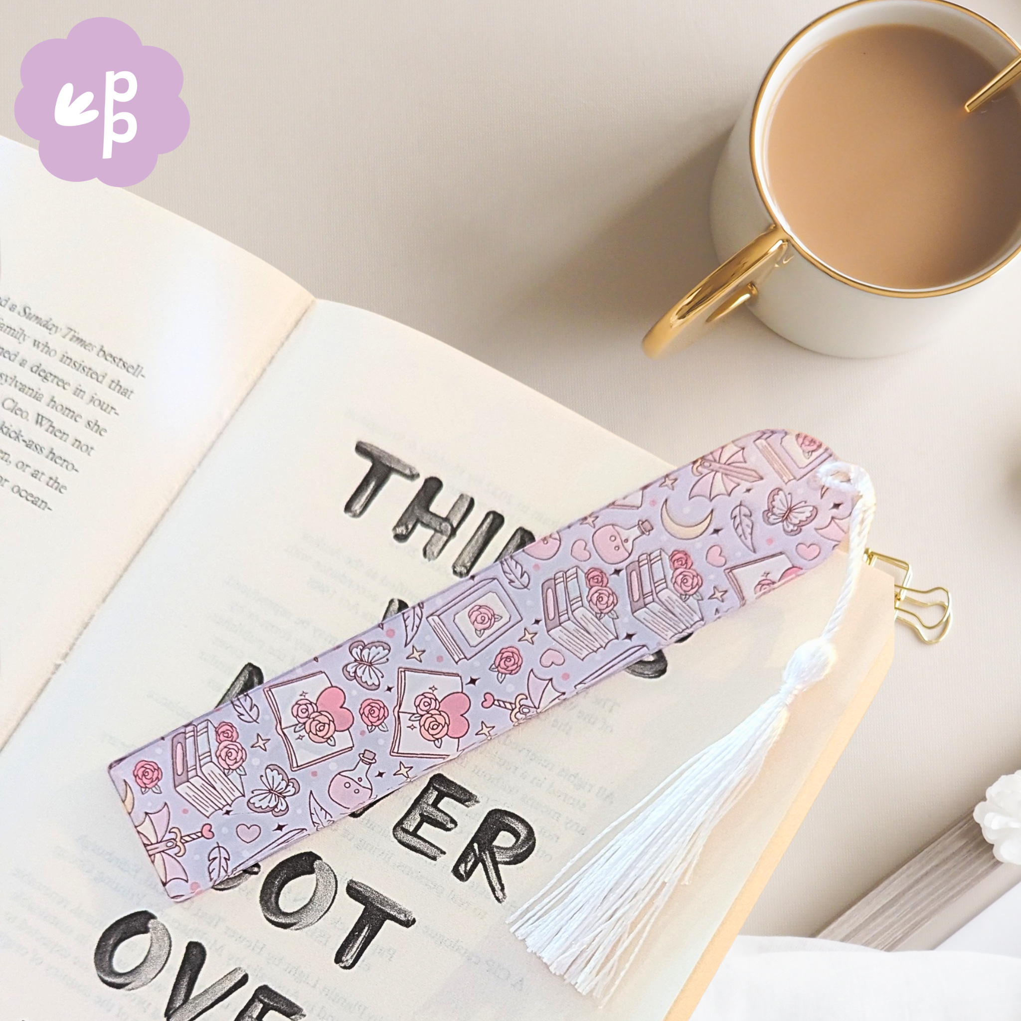 Handmade pastel acrylic bookmark with white tassel on open book next to a cup of tea.