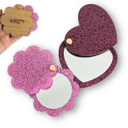 Two glittery compact mirrors, one heart-shaped and one flower-shaped, with a hand holding a wooden card in the background.