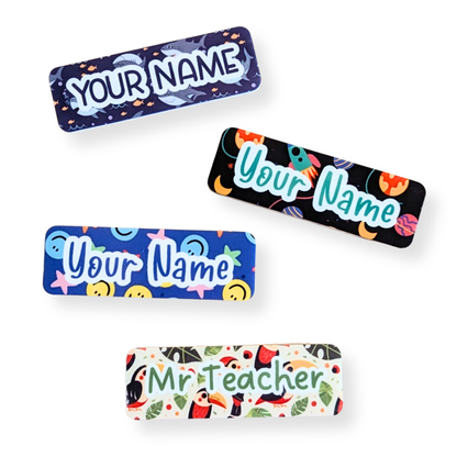Unisex teacher name badges featuring fun patterns like sharks, rockets, smiley faces, and tropical birds on magnetic bamboo backings – customizable by Priority Play.